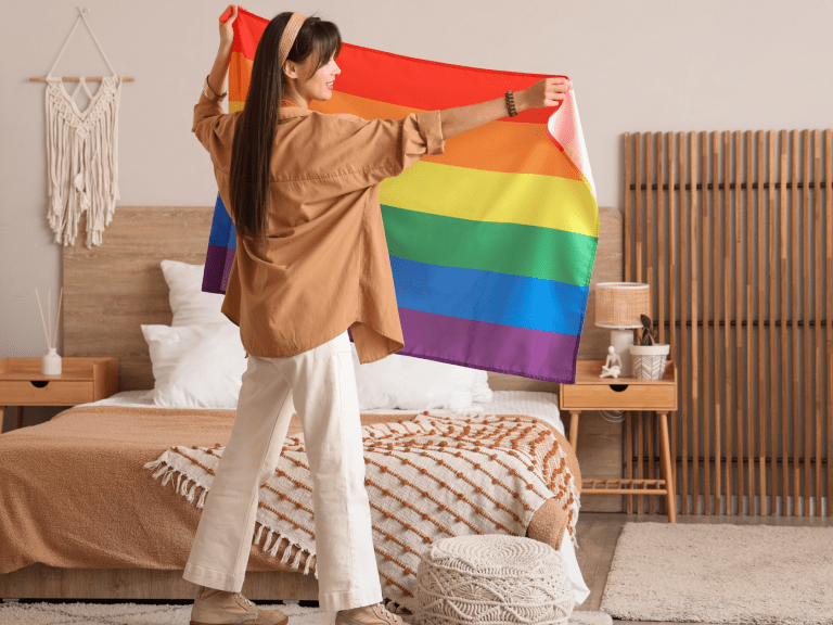 Person decorating a cozy bedroom by hanging a rainbow pride flag above a neatly made bed, symbolizing LGBTQ+ pride and home refresh on a budget.